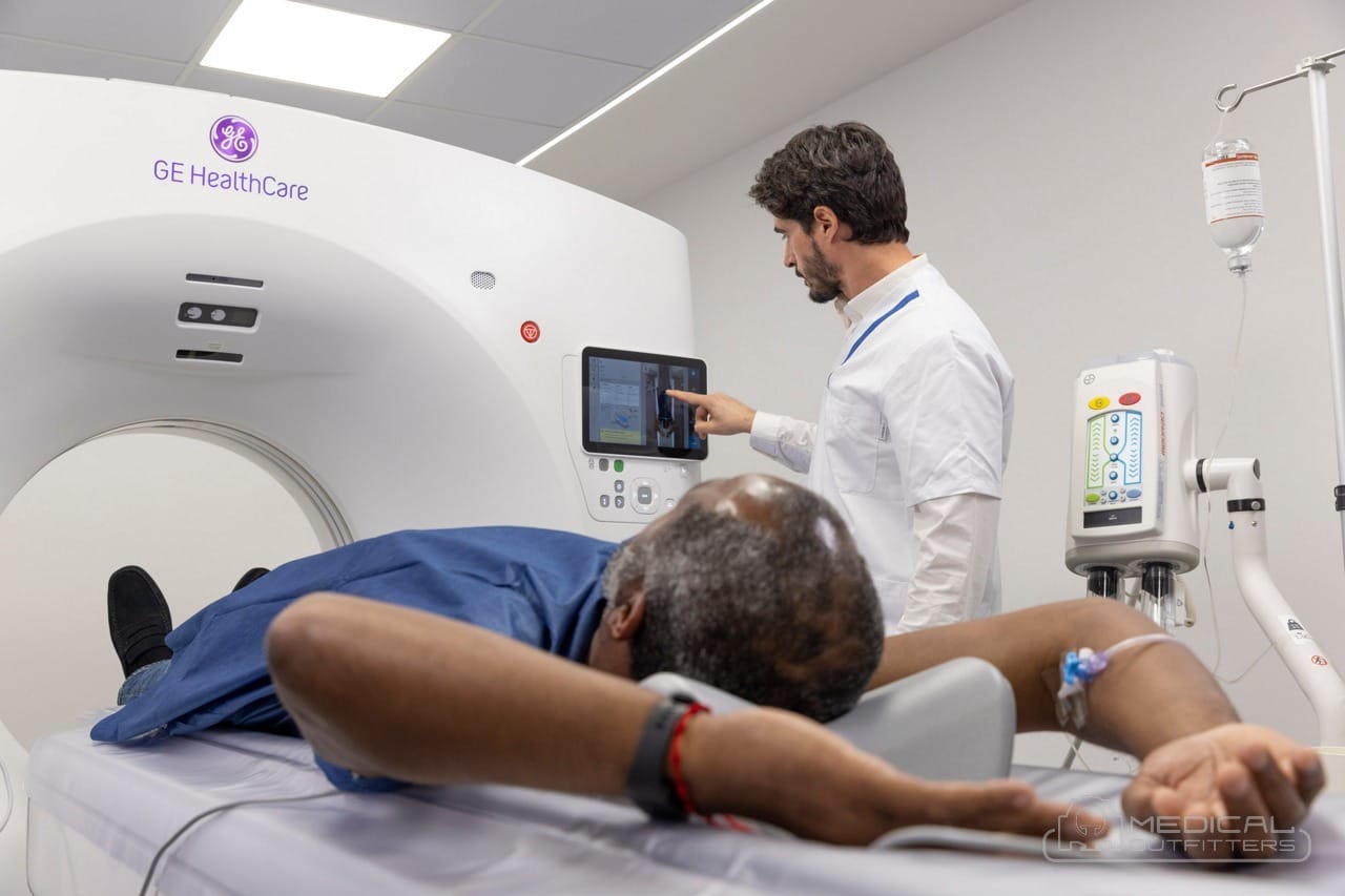 Technologist using the gantry touchscreen of a GE HealthCare CT scan machine while a patient lies on the table, photo courtesy of GE HealthCare